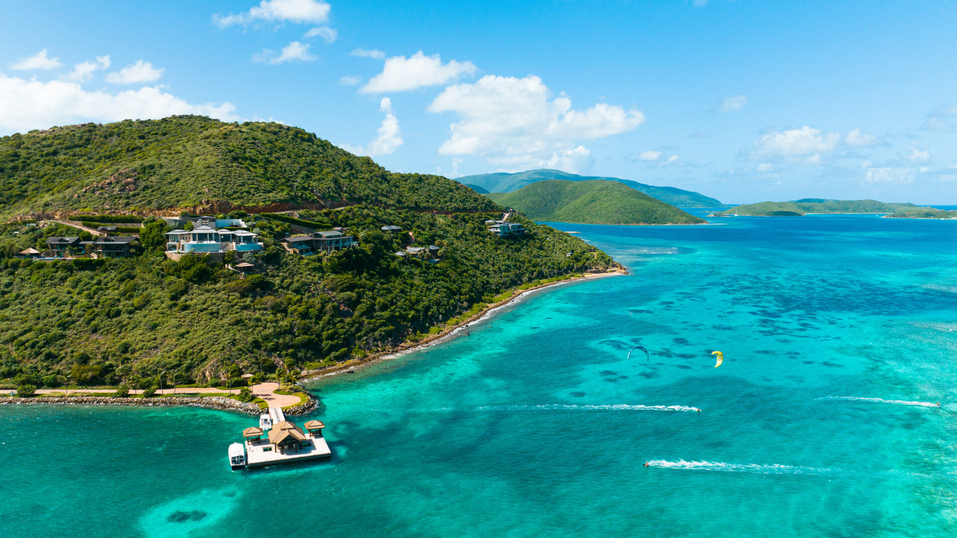 A lush green island surrounded by turquoise waters is shown. Several houses are perched on the hillside. In the foreground, a floating dock with a small bungalow is visible, and a kite surfer glides over the water nearby. Celebrating World Oceans Day, this idyllic retreat highlights premier resorts under bright skies with scattered clouds.