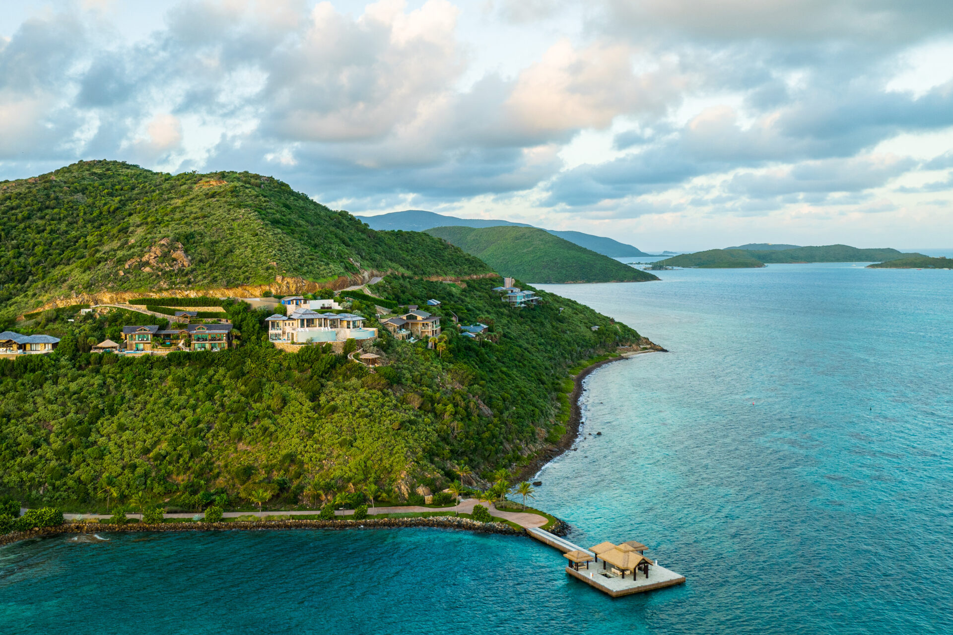 Aerial view of lush green hills by the ocean with several luxury houses nestled on a promontory. The foreground features a wooden pier and small dock structure over clear blue waters. The sky is partly cloudy, and distant islands are visible on the horizon—one of the best places to travel in April.