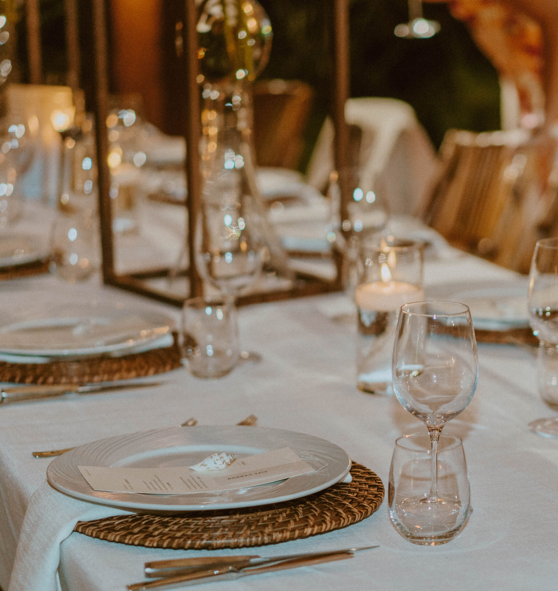 A sophisticated dining table by BVI Weddings & Events is set for a formal event with white tablecloths, wicker placemats, neatly arranged plates, menus, silver cutlery, wine glasses, and water glasses. Candles and floral arrangements in tall, slender vases add an elegant touch.