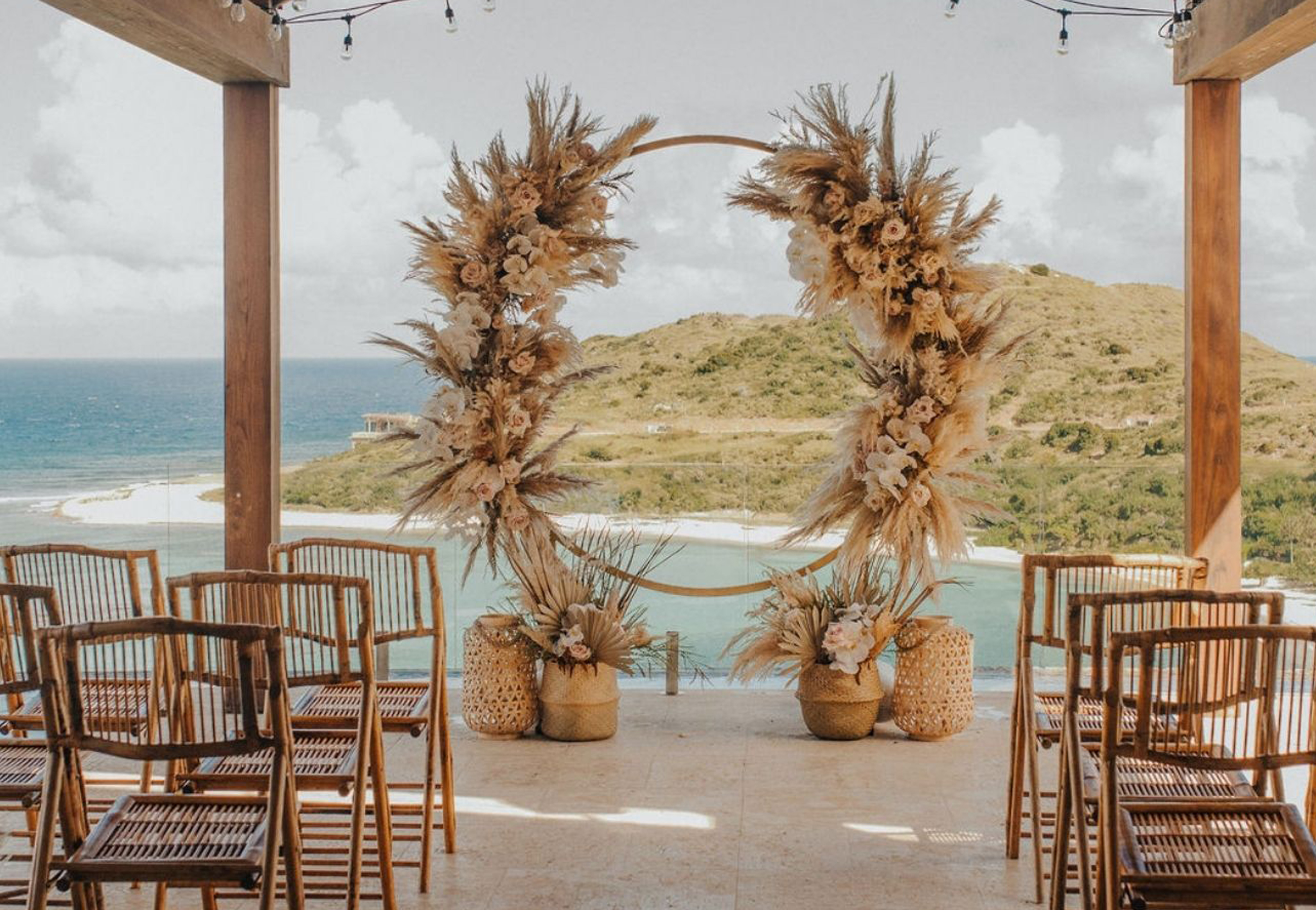 A wedding altar setup by BVI Weddings & Events features a large circular floral arrangement made of dried flowers and grasses. The altar overlooks a scenic view of the ocean and a green hillside. Wooden chairs are arranged in rows, with string lights delicately strung above.