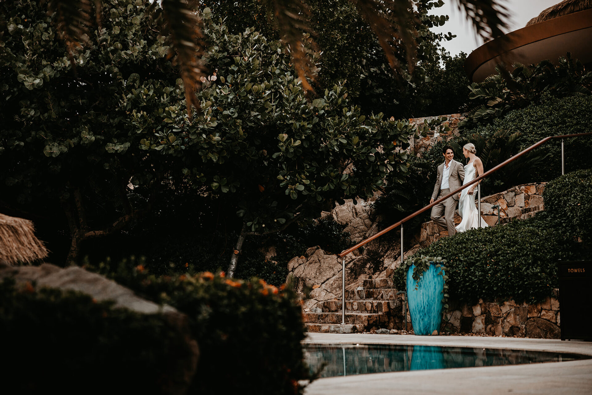 A couple dressed in wedding attire descends a stone staircase surrounded by lush greenery and rock formations. Smiling and holding hands, they approach a clear blue swimming pool. The serene and intimate scene, orchestrated by BVI Weddings & Events, is set in a tropical outdoor paradise.