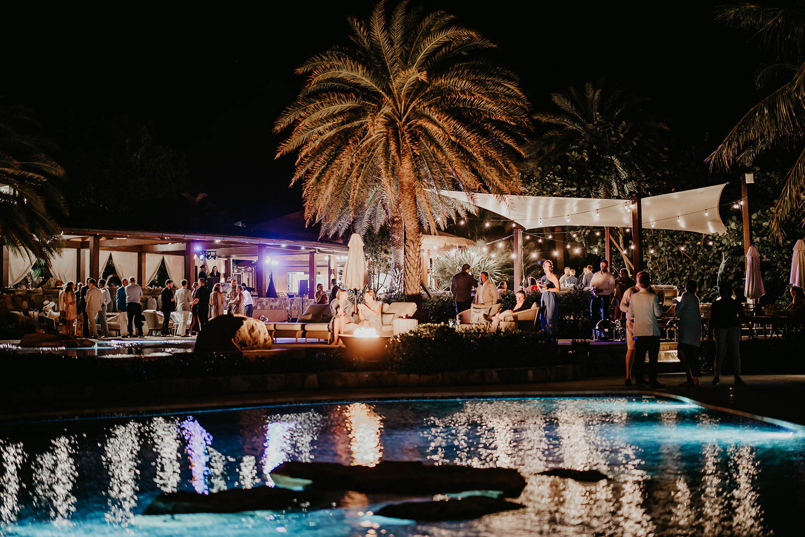 A nighttime outdoor party by a pool, hosted by BVI Weddings & Events, features people socializing under elegant lighting and amidst palm trees. Live music plays on a small stage, contributing to the lively atmosphere, while reflections shimmer in the illuminated pool water.