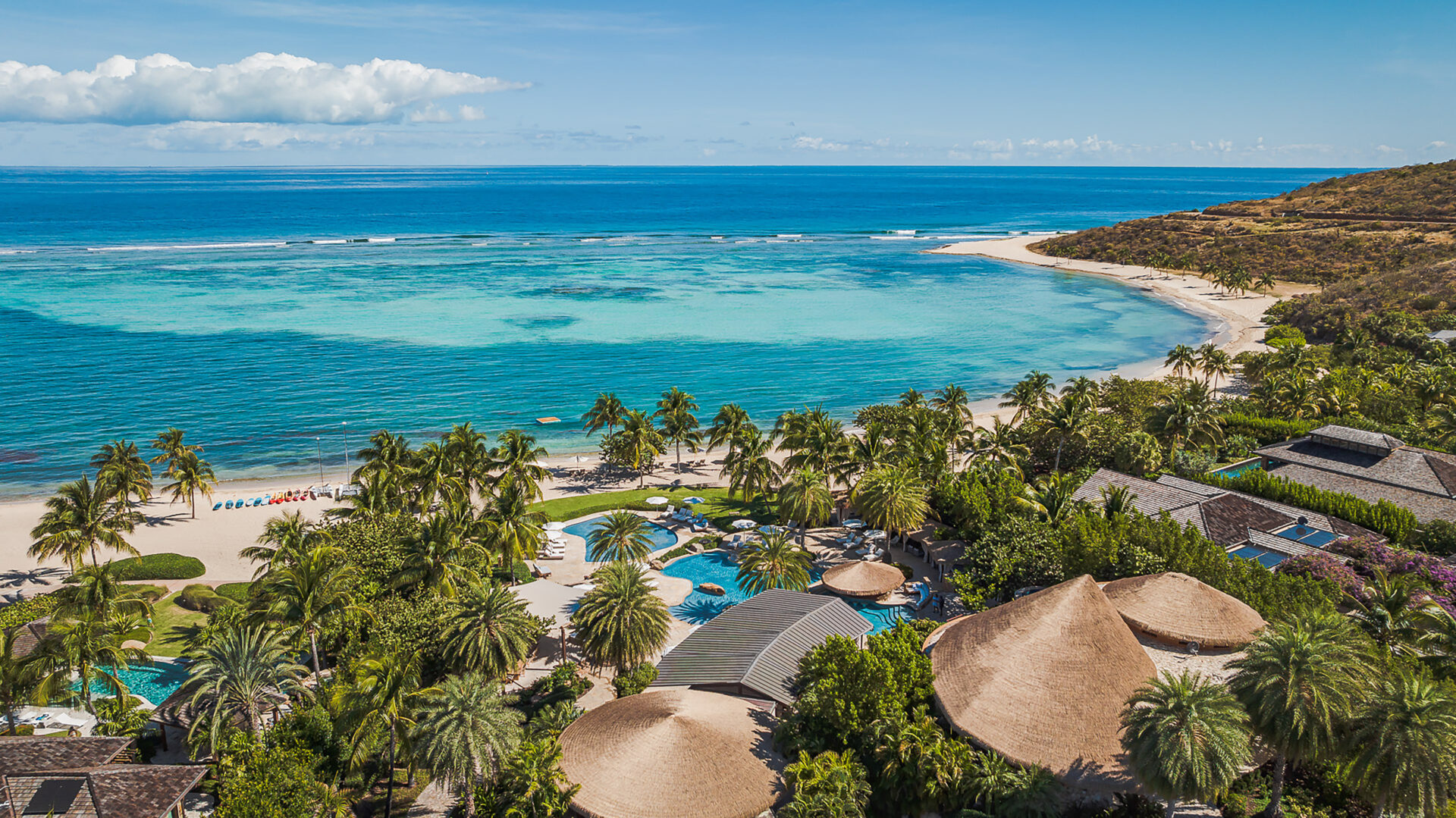 Aerial view of a tropical beach resort featuring several thatched-roof buildings, lush green palm trees, and multiple swimming pools. Nestled in the heart of the hottest Caribbean property market, the resort overlooks a stunning blue-green ocean and a sandy beach with sun loungers.