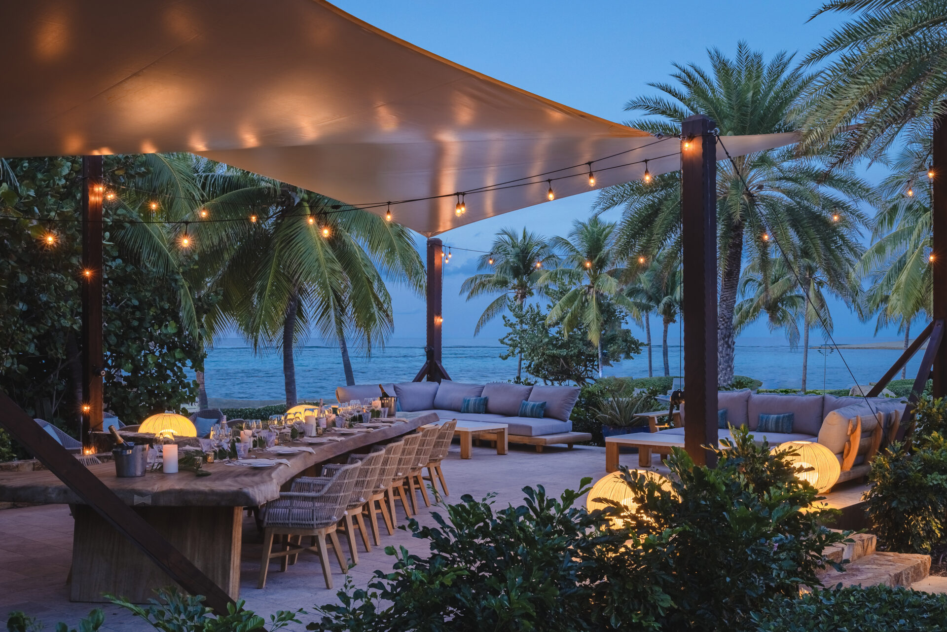 An elegant outdoor dining area by the sea, featuring a long wooden table set with candles, wicker chairs, and vibrant greenery. Overhead, string lights and a canopy create a cozy ambiance. Palm trees and a calm ocean at Oil Nut Bay can be seen in the background, offering an idyllic spring break escape during twilight.