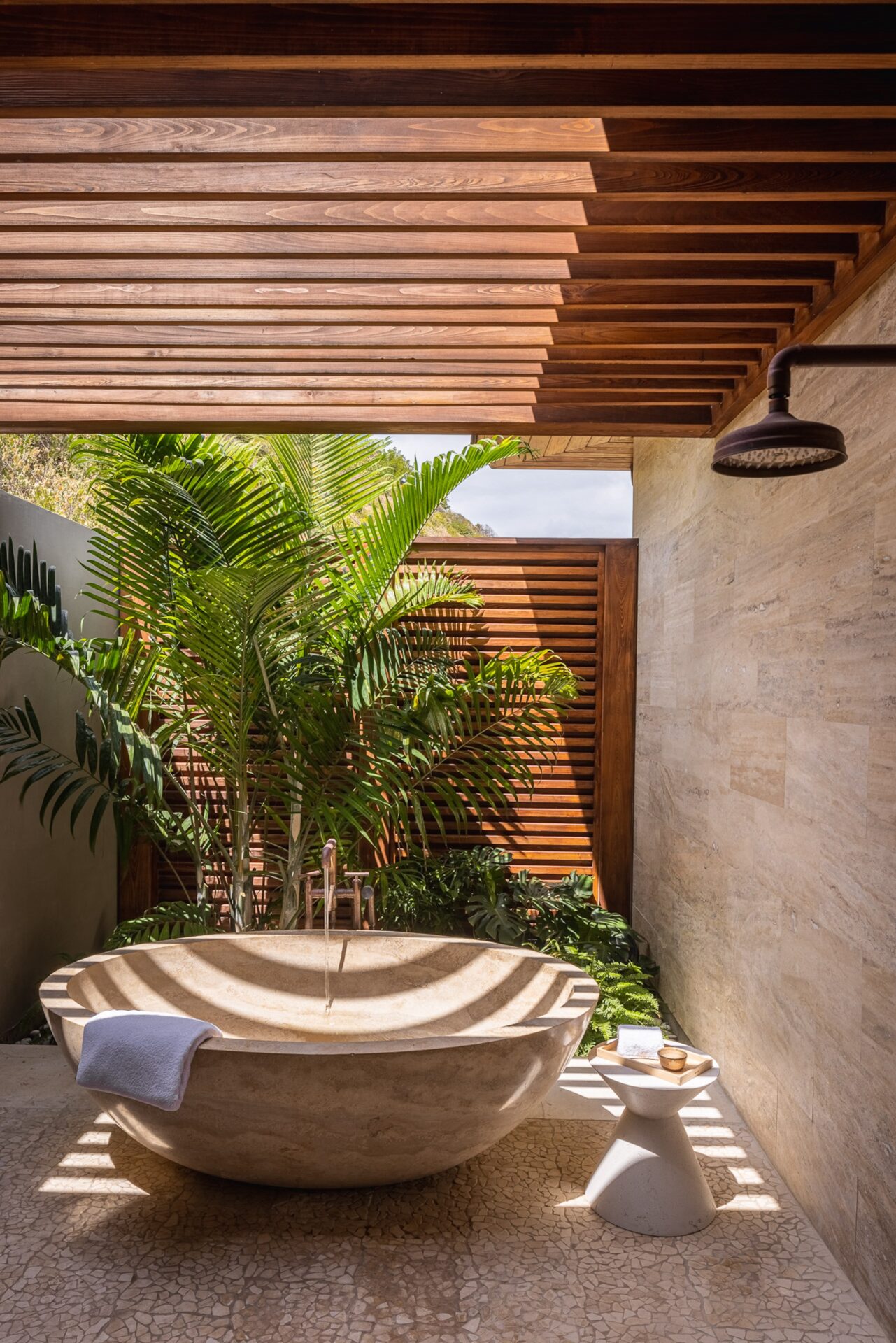 A luxurious outdoor bathroom at Xela Villa features a large stone bathtub under a pergola with wooden slats. Lush green foliage surrounds the space, and there is a small white stool with a towel and toiletries next to the tub. Located in the beach neighborhood, the area is bathed in natural light.