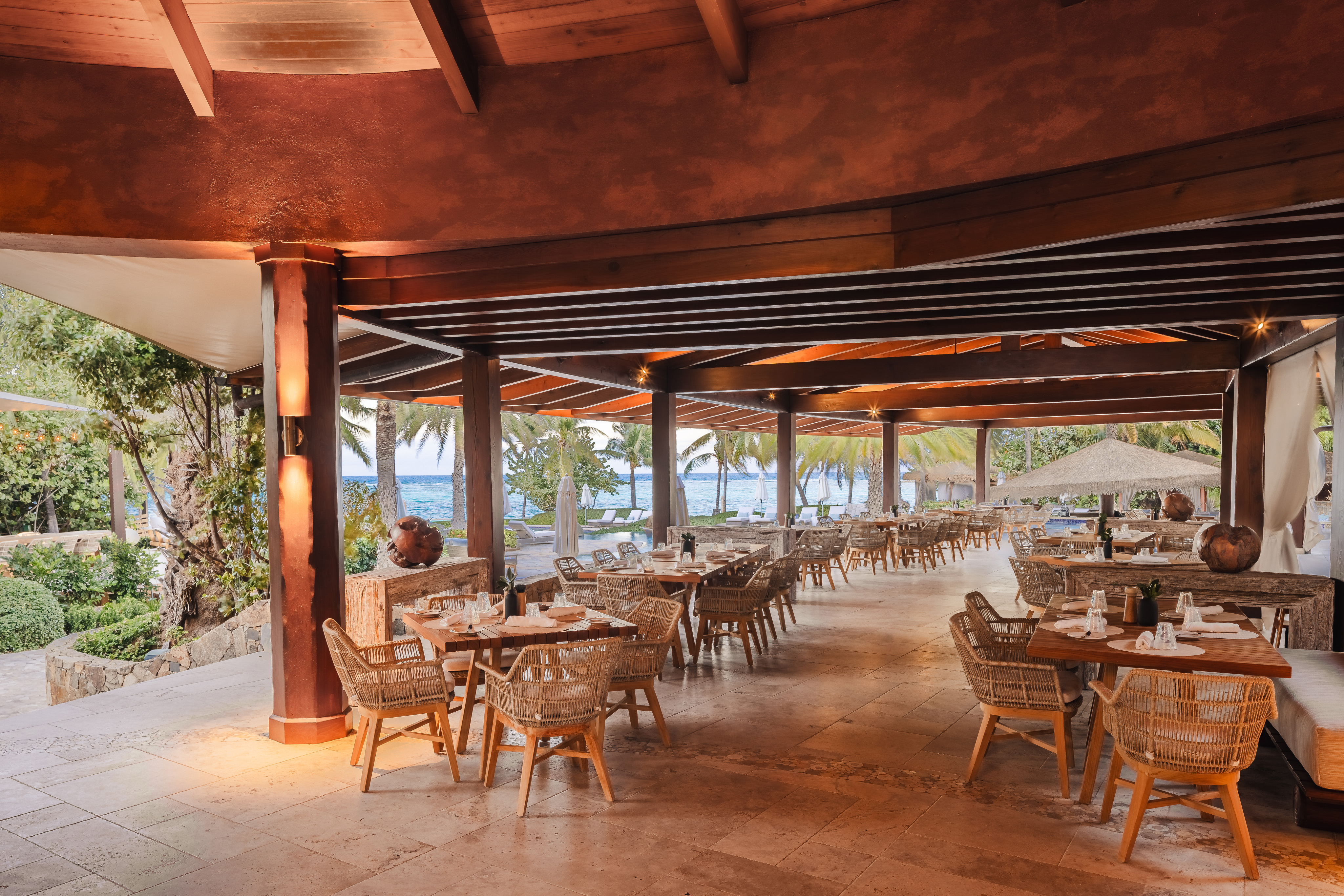 An open-air restaurant with a wooden roof and pillars features tables with rattan chairs. The seating area extends to a lush garden with palm trees, and beyond that, a seaside view is visible. Island dining in the British Virgin Islands embraces natural light, creating a relaxed and airy atmosphere.