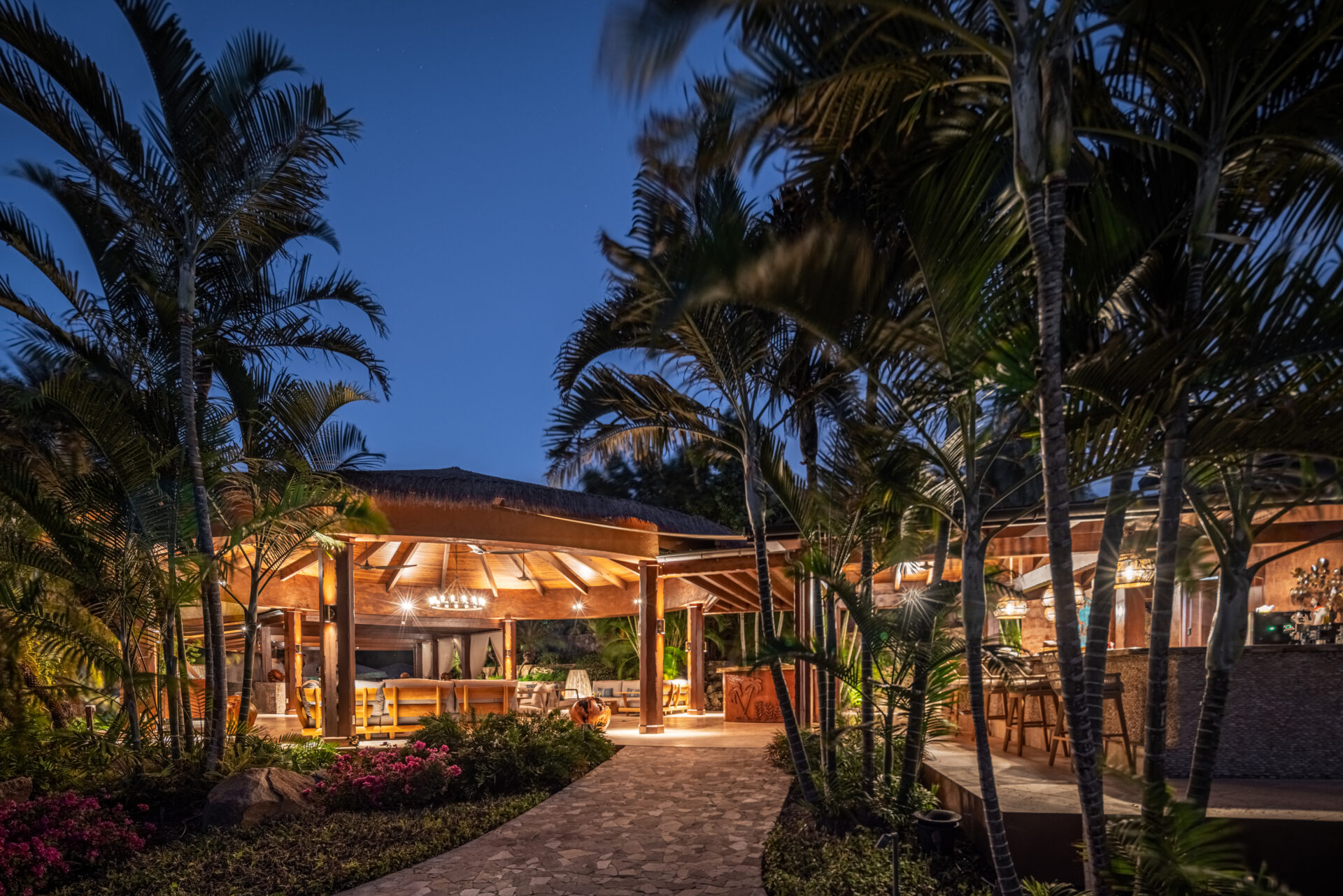 A scenic view of a tropical outdoor dining area during the evening, offering island dining in the British Virgin Islands. The space is beautifully lit with chandeliers and surrounded by lush palm trees and vibrant greenery. A stone pathway leads to the dining space, creating a welcoming and serene atmosphere.