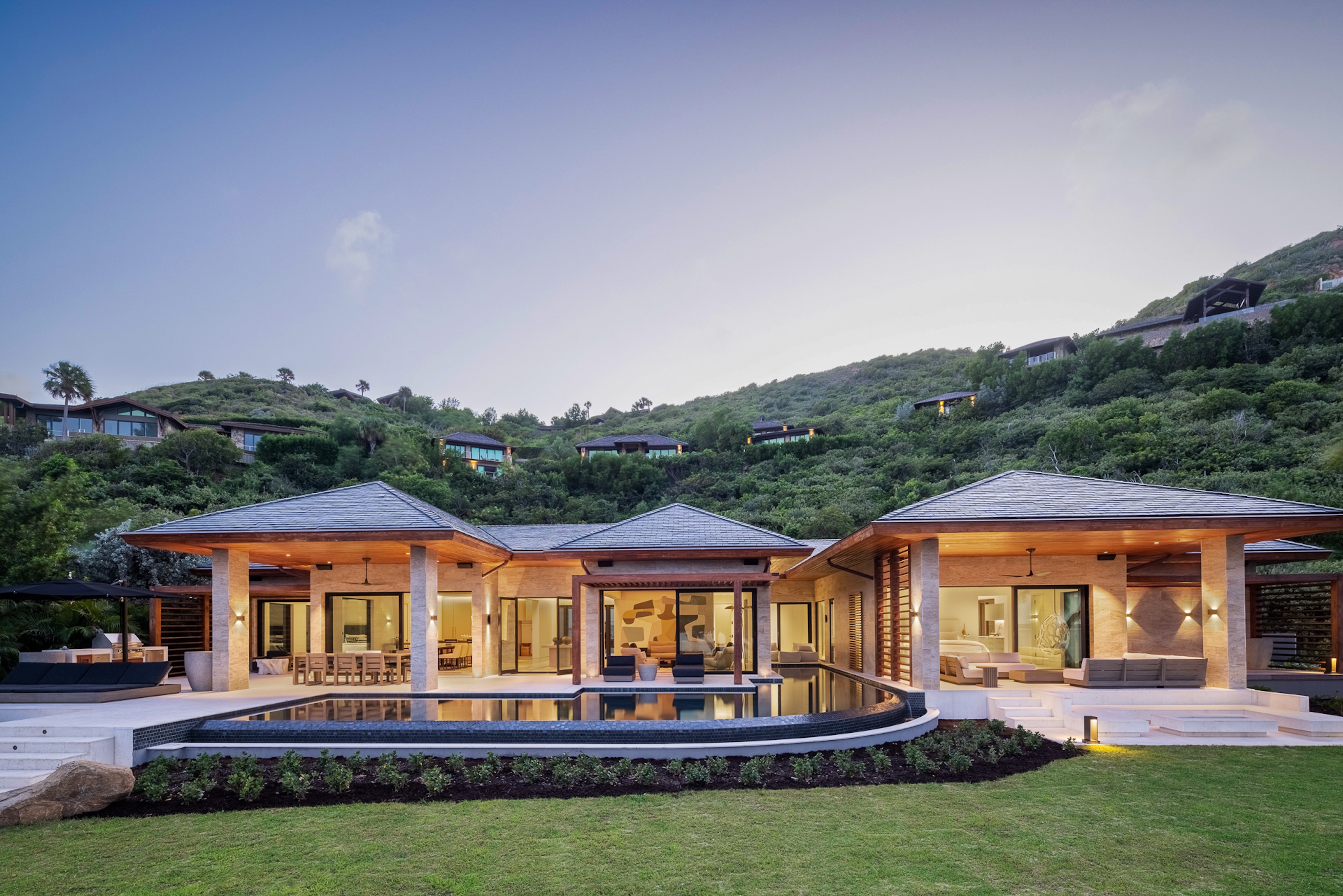 Modern, luxurious Xela Villa with three connected sections, large glass windows, a sloped gray roof, and a spacious pool in the foreground. The house is set against a lush, green hillside dotted with other residences in an exclusive beach neighborhood, under a clear evening sky.