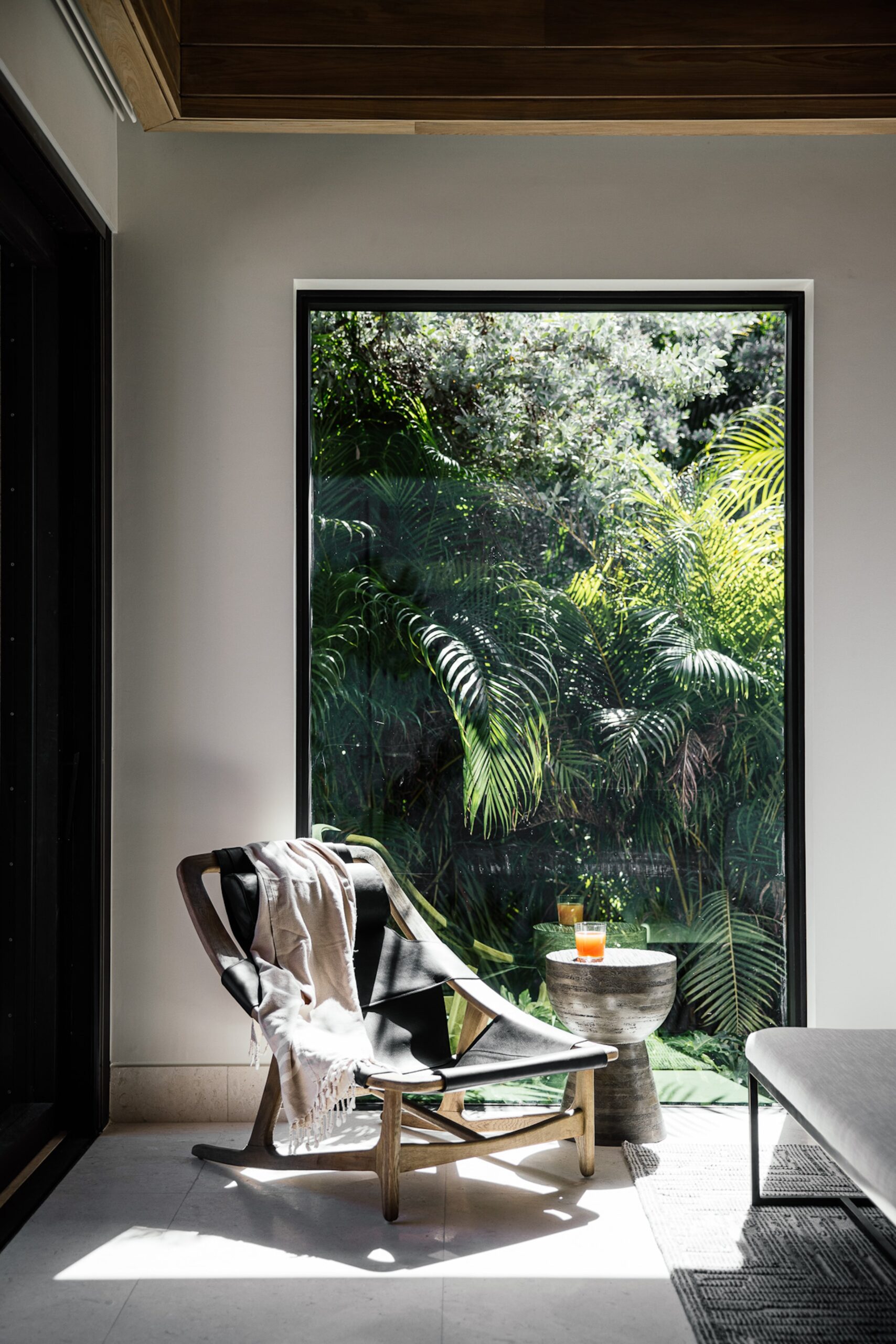 A cozy reading nook featuring a modern wooden lounge chair with a draped blanket and a small side table holding a drink. The space is bathed in natural light from a large window, offering a lush view of tropical plants outside, perfect for unwinding in the serene neighborhood of Xela Villa.