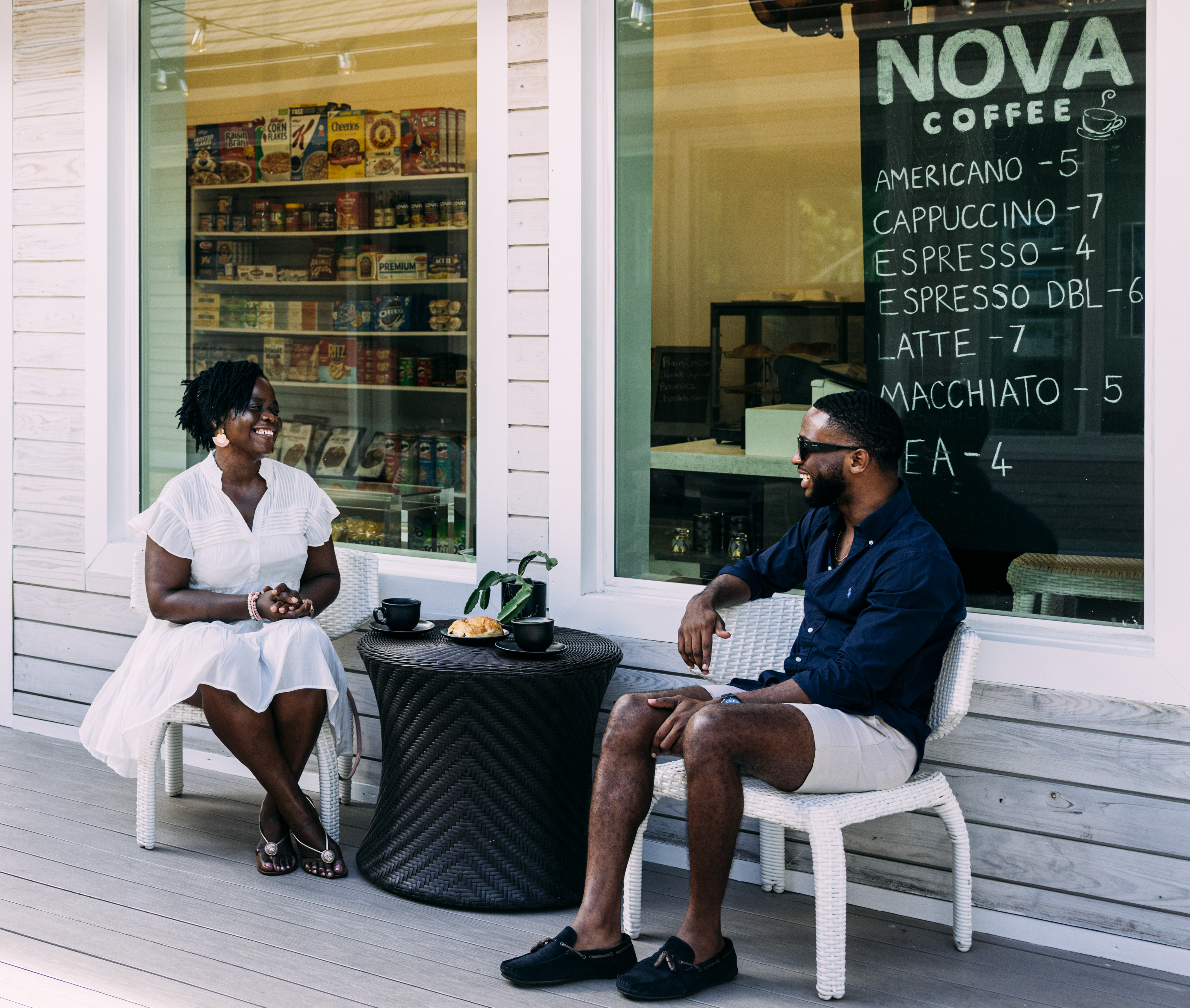 A woman and a man are sitting outside Nova Coffee at Marina Village in the British Virgin Islands. They are smiling and enjoying coffee at a small table. The café window displays various prices. The woman is in a white dress, while the man sports a dark shirt and shorts.