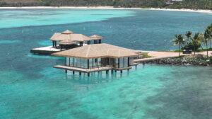 A luxurious overwater bungalow with a thatched roof sits above turquoise waters, soon to host the exclusive Sundara Spa Studio. The structure features large windows and is connected to the land by a walkway. Nearby, another similar bungalow and some palm trees are visible in the background.