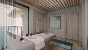 A serene overwater spa room with an ocean view features a massage table adorned with towels and purple flowers. The minimalist design includes a large window, light wood accents, and a scenic balcony overlooking the British Virgin Islands' shimmering waters.