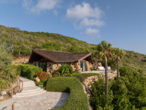 A rustic stone cottage with a wooden roof is nestled into a lush hillside on the Virgin Gorda Estate. Bright green plants and colorful flowers surround it. A winding stone path leads to the cottage, past a serene pool, with palm trees and a clear blue sky in the background.