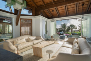 Luxurious open-concept living room of this $32.5 million estate on Virgin Gorda, with two beige sofas, a wooden coffee table, and blue armchairs. The space boasts a high wooden ceiling, modern lighting, and large windows that overlook palm trees and a lush garden.