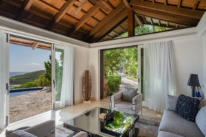 A cozy living room with light-colored sofas and a glass coffee table opens up to scenic views at the $32.5 Million Virgin Gorda Estate. Two large sliding doors reveal a shimmering pool and lush greenery outside, while the wooden ceiling adds warmth to this luxurious space.