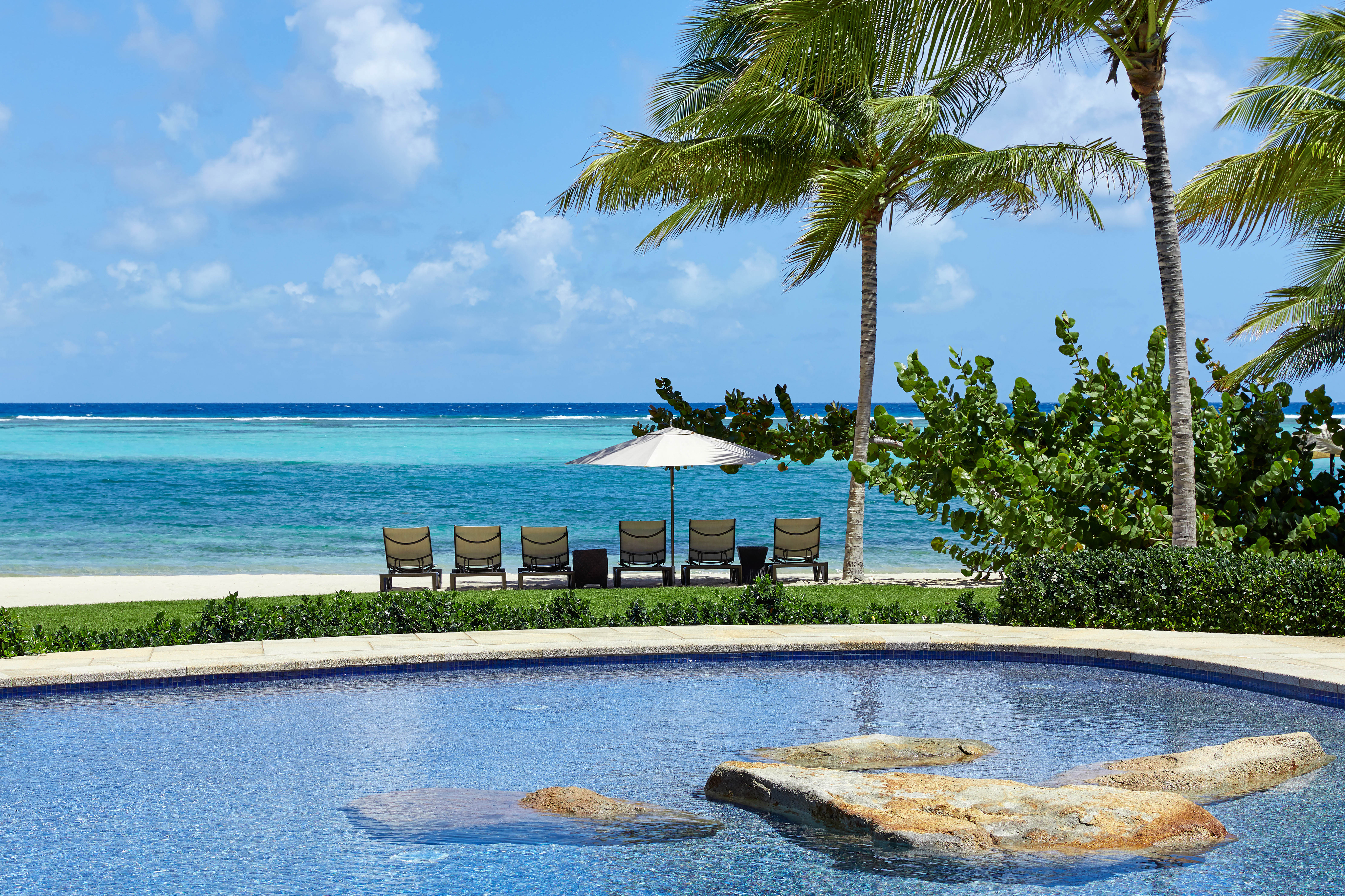 Enjoy a serene beachfront view perfect for getaways, with a shimmering pool in the foreground. Lounge chairs and a large umbrella await by the seaside, facing turquoise waters. Palm trees sway gently under a bright sky with scattered clouds—ideal for catching last-minute deals or planning Valentine's Day retreats.