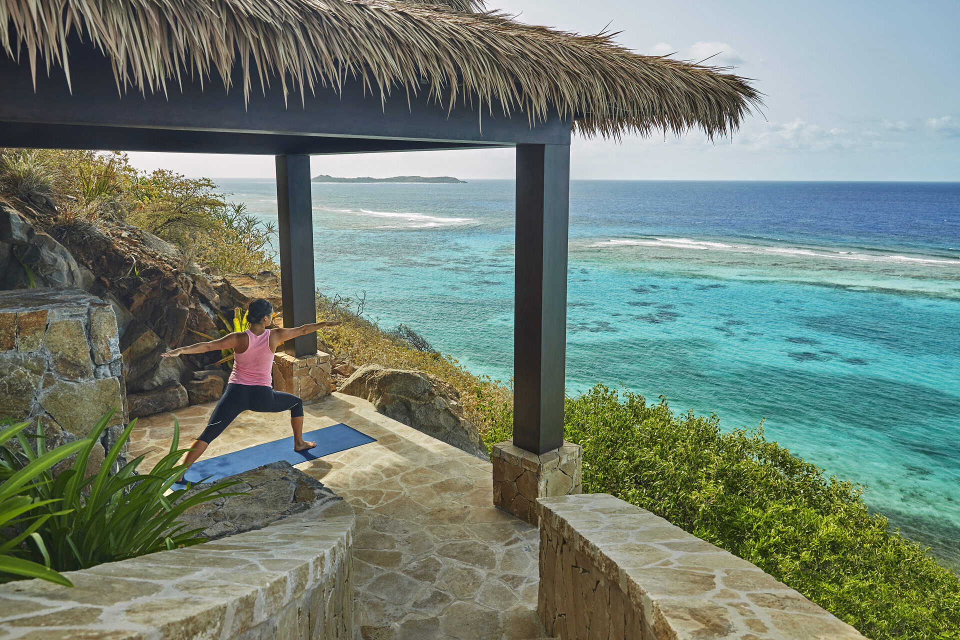 A person practices yoga on a mat under a thatched canopy at a serene resort overlooking the turquoise ocean. Embracing the Blue Mind Theory, stone steps and lush greenery surround them, with a clear blue sky in the background.