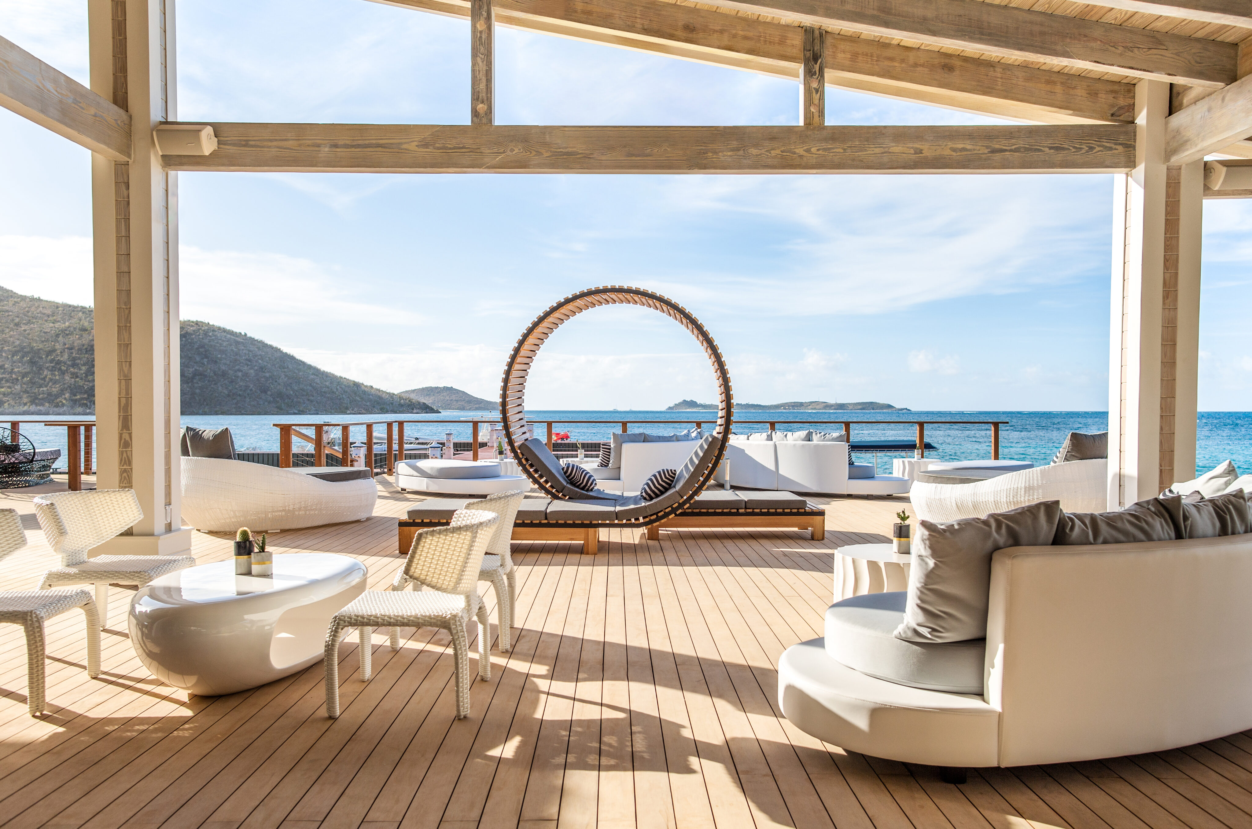 This luxurious outdoor lounge, one of the best places to unwind, features modern white furniture and a circular canopy daybed on a wooden deck overlooking the ocean. With scenic views of hills and blue sky in April, it creates a tranquil and inviting atmosphere.