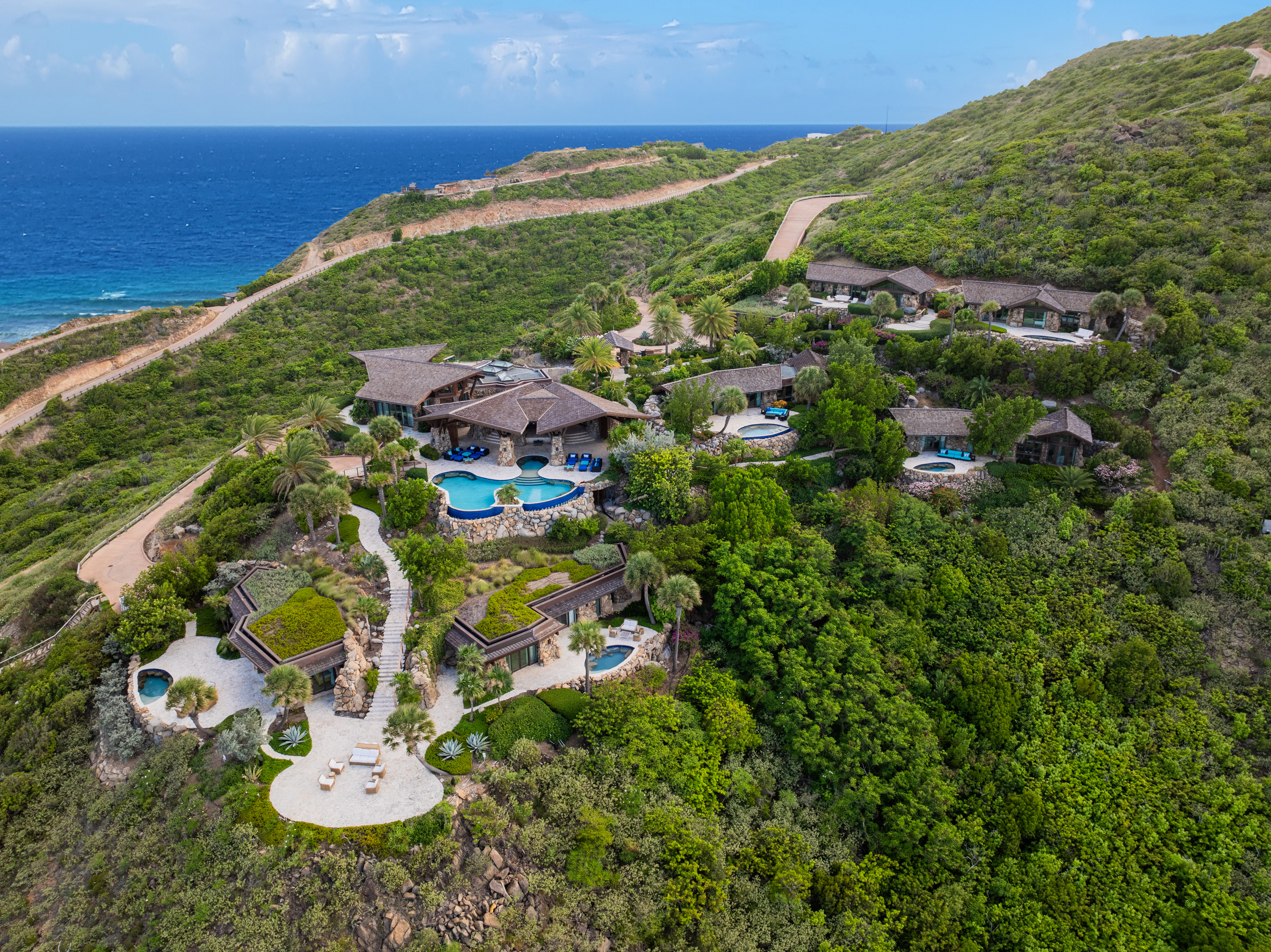 Aerial view of a lush green hillside with a luxurious villa complex at Oil Nut Bay. The design-forward villas feature thatched-roof buildings, circular pools, and winding pathways, all overlooking the vibrant blue ocean under a clear sky.